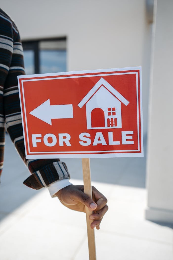 gallery-3 Close-up of a hand holding a For Sale real estate sign with a house icon outdoors.